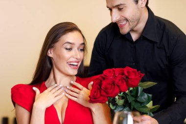 Making Surprise. Excited beautiful girlfriend in dress receiving bouquet of red roses from her happy handsome boyfriend who holding bunch of flowers during romantic dinner in luxury restaurant or cafe