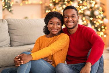Eğlence ve aile tatilinin evde kutlanması. Gülümseyen bin yıllık Afro-Amerikan erkek ve kadın rahat oturma odasında kameraya sarılıyor ve ona bakıyor.
