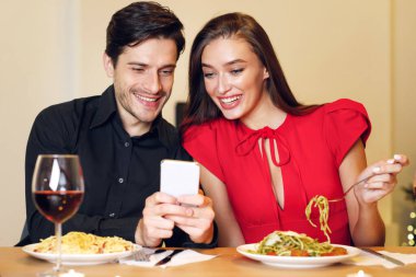 Happy Loving Family. Beautiful young millennial couple using cellphone, sitting at table and having dinner, eating pasta. Smiling man sharing photos with his excited woman, spending time together