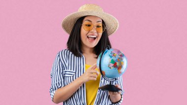 Maceraya hazırım. Heyecanlı, mutlu Asyalı bir kadının dünya küresi tutan portresi, haritaya ve planlı tatile işaret eden, gideceği yeri seçen, gözlük takan ve yaz hasır şapkası takan, pembe stüdyo geçmişi olan