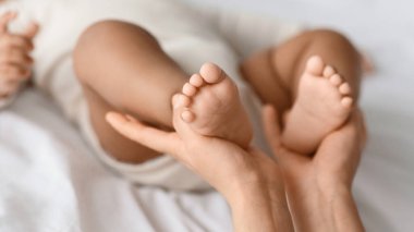 Anne sevgisi kavramı. Küçük Afrikalı Amerikalı bebeğini elinde tutan genç anneye yakın çekim. Evde beyaz battaniye örtüsüne uzanmış bebek kıyafeti giymiş, bacaklara odaklanmış.