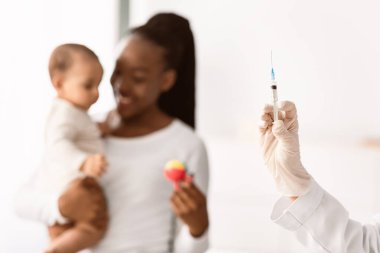 Pediyatrik konsept. Doktorun şırıngayı tutmasına, gülümseyen siyahi bir annenin bebeğini eline alıp ona oyuncağını göstermesine odaklan. Çocuk aşısı, hastanede sağlık aşısı.