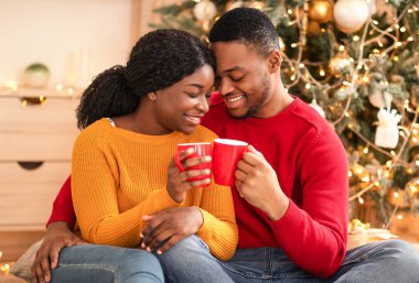 Christmas mood, free time and celebrate New Year together. Smiling young african american couple clink cups with cocoa, sits on floor and hugging in home interior with tree with glowing garlands