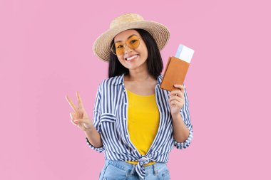 İyi yolculuklar. Pasaport ve bilet tutan neşeli Asyalı kadın portresi, barış işareti işareti taşıyan, pembe stüdyo arka planında zarif hasır şapka ve güneş gözlüğü takan pozitif turist kadın.