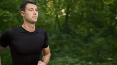 Genç bir adam yemyeşil bir parkta dışarıda koşuyor, sıcak yaz gününün tadını çıkarıyor. Sağlıklı bir yaşam tarzını geliştirmek için spor rutinini uygularken odaklanmış bir ifade takınıyor..