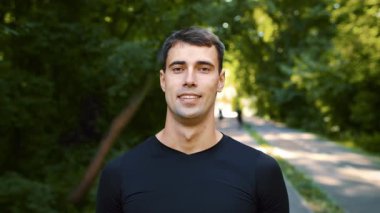 Spor kıyafeti giymiş genç bir adam parkta duruyor, koşudan sonra gülümsüyor. Bereketli yeşillik onu çevrelemiş, sıcak yaz mevsiminde canlı ve aktif bir yaşam tarzını yansıtmaktadır..
