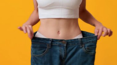 Genç bir kadın büyük beden kot pantolon tutarak kilo verme yolculuğunu sergiliyor. Parlak sarı arkaplan, onun başarısını ve kendine güvenini neşeli bir ortamda vurguluyor..