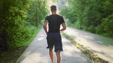 Genç bir adam parkta ağaçlarla kaplı bir patika boyunca koşuyor, temiz havanın ve güneşin tadını çıkarıyor. Sıcak bir günde çevresi doğa ile çevriliyken aktif bir yaşam tarzını benimsiyor..