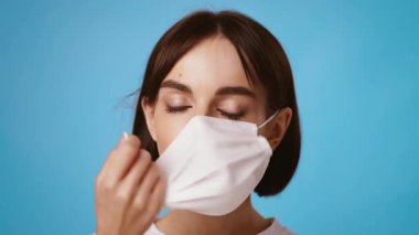 Genç esmer bir kadın gözleri kapalı, beyaz bir maske takıyor. Sakince maskesini ayarlarken sakin atmosfer ondan yayılıyor. Arka plan rahatlatıcı bir mavi..