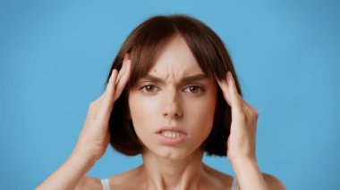 Genç bir esmer kadın, iki eliyle başını tutarken gözle görülür şekilde hayal kırıklığına uğrar. Katı mavi arka plan ifadesini güçlendiriyor, yoğun bir duygu hissi veriyor. Migren, baş ağrısı