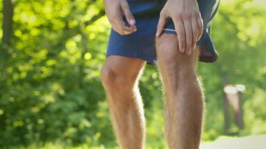 Genç bir adam yaz boyunca parkta koşuyor. Yeşilliklerle çevrili dizindeki rahatsızlığı değerlendirmek için duruyor. Sağlıklı yaşam tarzını ve fitness hedeflerini vurguluyor..
