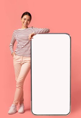 Mobil Teklif. Güzel, gülümseyen Asyalı bir kadın boş beyaz ekranlı büyük akıllı telefona yaslanıyor tasarımınız ya da reklamınız için bedava fotokopi alanı gösteriyor, pembe arka plan üzerinde poz veriyor,