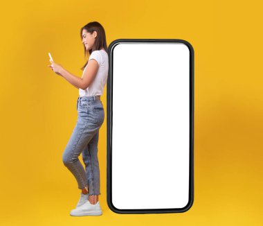 Harika bir uygulama. Beyaz ekranlı ve cep telefonu kullanan mutlu bayan, sosyal medyada neşeli kadın sohbeti, turuncu arka planda durmak, görüntüyü taklit etmek, vücut uzunluğu...