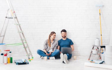 Ev geliştirme veya onarım sırasında dizüstü bilgisayar kullanan genç çift, beyaz tuğla duvar arka planı, kopyalama alanı