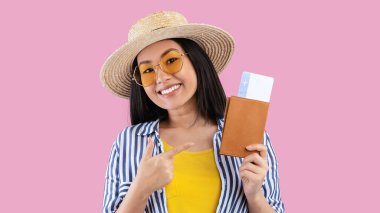 Tatil zamanı. Pasaport ve uçak biletlerini tutan heyecanlı Asyalı kadın yaz tatili için hazır, hasır şapka ve güneş gözlüğü takıyor, pembe stüdyo duvarında izole edilmiş,