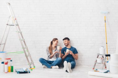 Yenileme, onarım ve aile kavramı. Genç bir çift yeni evinde tamirat yapıyor ve kahve içip rahatlıyor.