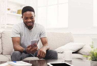 Sosyal ağlar. Evde Smartphone'da mesajlaşma kulaklık genç mutlu Afrikalı-Amerikalı adam. Müzik, bej koltukta oturan, alanı Kopyala