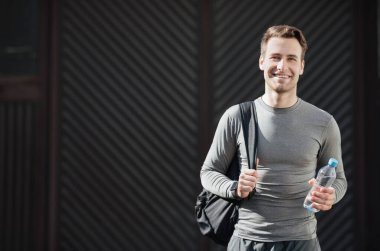 Elinde su şişesi olan adam eğitime hazır. Spor kıyafetleriyle gülümseyen sporcu, spor salonuna gider. Duvarın arkasında, boş alanda.
