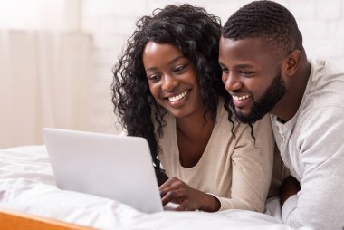 Mutlu Afro-Amerikan çift birlikte yatarken dizüstü bilgisayar kullanıyor, internette geziniyor ya da film seyrediyor.