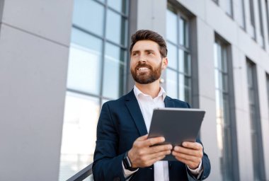 Sakallı, tablet yazan mutlu, kendine güvenen, yakışıklı beyaz adam ofis binasının yakınındaki boş alana bak. Açık havada başarılı bir iş, iş adamı ticaret, reklam ve teklif için uygulama kullanıyor