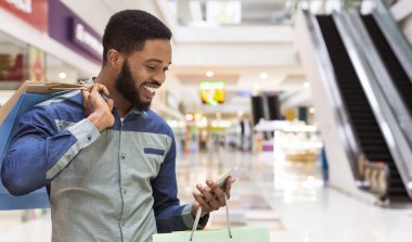 Cep telefonu kullanarak alışveriş çantaları ile yakışıklı Afrikalı Amerikalı adam ve Mall merkezinde gülümseyen, Panorama, kopya alanı