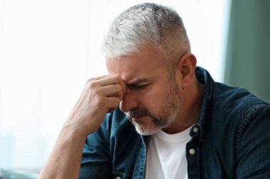 Bitkin, kır saçlı, sakallı bir adam burun köprüsüne dokunuyor, kanepeye oturuyor, gözleri kapalı, ev içi. Yaşlı adam kendini hasta hissediyor, baş ağrısı ya da akşamdan kalma, kopyalama alanı.