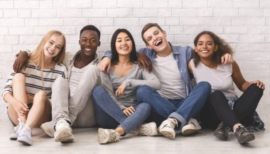 Zeminde oturan çok ırklı öğrenciler, beyaz tuğla duvarın üzerinde kucaklaşıp eğleniyorlar, kamera önünde poz veriyorlar.