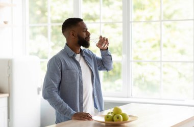 Sakin bin yıllık Afro-Amerikan sakallı adam pencerenin arkasında elmalarla mutfak içlerindeki camdan su içiyor. Susuzluk, su dengesi, sağlıklı beslenme ve su kaybı.