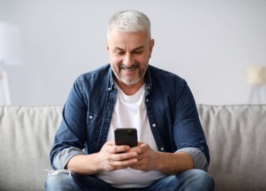 Gülümseyen, gri saçlı, koltukta oturan, cep telefonu kullanan, fotokopi çeken biri. Akıllı telefonlu neşeli orta yaşlı bir adam ailesiyle sohbet ediyor ya da mobil uygulama kullanıyor.