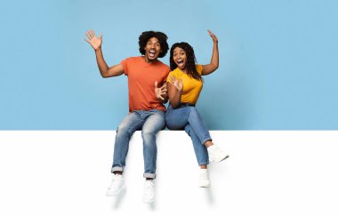 Heyecanlı siyahi aşıklar yatay beyaz tahtada oturup mesaj ya da reklam yazıyorlar. Mutlu Afro-Amerikan erkek ve kadınlar çığlık atıp el kol hareketi yapıyorlar. Mavi stüdyo arka planı, kopyalama alanı.