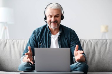 Başarılı yakışıklı, sakallı, olgun bir adam evden video iş görüşmesi yapıyor, kulaklık ve laptop kullanıyor, oturma odasında koltukta oturuyor, iş arkadaşlarıyla konuşuyor, gülümsüyor ve el kol hareketi yapıyor, uzay kopyaları yapıyor.