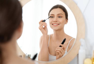 Gülümseyen genç bir kadın makyaj yapıyor, aynaya bakıyor, maskara kullanıyor, ev dekorasyonu yapıyor.