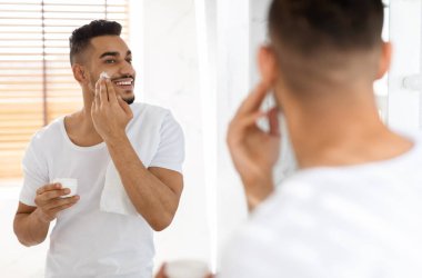 Erkek Güzellik Hizmetleri. Yakışıklı genç Arap adam banyoda nemlendirici yüz kremi sürüyor, aynaya bakan gülümseyen Orta Doğulu adam ve cildine krem sürüyor,