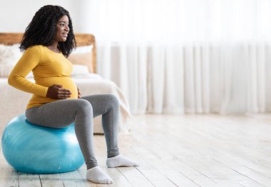 Koca göbekli, gülümseyen Afrikalı hamile kadın yatak odasında spor salonunda oturup fotokopi odasına bakıyor. Hamilelik sırasındaki spor. Siyahi kadın baloda antrenman bekliyor, doğum için hazırlanıyor.