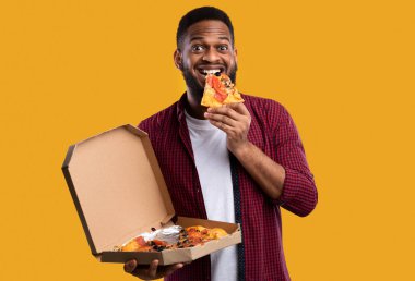 Kameraya gülümseyen, sarı arka planda duran, pizza yiyen mutlu Afrikalı bir adam. Abur cubur yiyici sağlıksız pizzanın tadını çıkarıyor. Beslenme ve aldatma yemeği, fast food yeme alışkanlığı