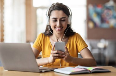 Kulaklık takmış genç bir kız, kafede oturuyor, dizüstü bilgisayar ve akıllı telefon kullanıyor.