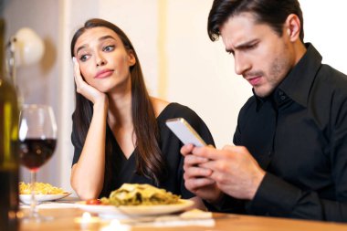 Bad Boring Date. Unhappy Sad Lady Sitting On Date Evening In Restaurant While Focused Man Using Cellphone Gadget And Texting Sms, Ignoring His Girlfriend. Relationship Problem Concept
