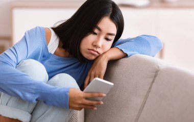 Depresyon Konsepti. Akıllı telefona bakan, sosyal medyayı kontrol eden, kanepede uzanan hayal kırıklığına uğramış Asyalı genç kadının portresi. Üzgün bayan mesajını ya da aramasını bekliyor, kötü haber okuyor.