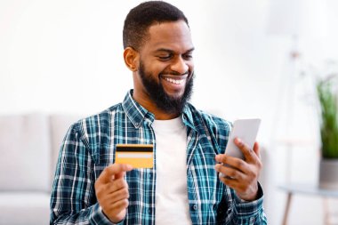 Evdeki koltukta oturan Smartphone üzerinden alışveriş yapan siyah adam kredi kartı tutuyor. Alıcı Bir şeyler satın alıyor Uygulamayı kullanarak. E-Ticaret, Mobil Bankacılık Uygulaması Konsepti. Seçici Odaklanma