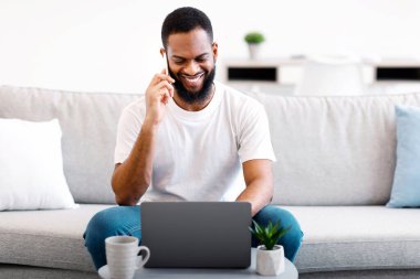 Laptop 'ta cep telefonuyla konuşan ve evde oturan mutlu siyah serbest çalışan adam. Uzaktan Çalışma ve Uzaktan Telefon İletişimi. Her zaman temastayım. Seçici Odaklanma