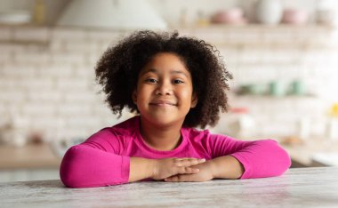 Mutfakta masada oturan kıvırcık siyah kızın yakın plan portresi, evde dinlenen Afro-Amerikan kadın çocuk, kreş çocuğu kameraya bakıyor ve gülümsüyor.