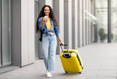 Neşeli, kıvırcık, orta doğulu, rahat kıyafetli genç bir kadın tek başına tatile çıkıyor, havaalanının yanından sarı bavulla geçiyor, cep telefonu kullanıyor, internetten kayıt yaptırıyor, fotokopi çekiyor, tam boy.