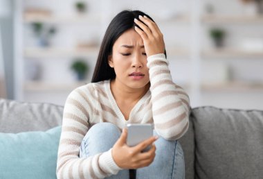 Endişeli Asyalı kadın akıllı telefon ekranına bakıyor ve kafaya dokunuyor, evdeki koltukta oturuyor, rahatsız Koreli genç bayan nahoş mesajlar okuyor, duygusal olarak kötü mesaja tepki veriyor, boş alan.