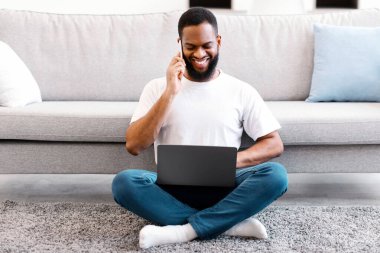 Afrikalı serbest çalışan bir adam dizüstü bilgisayar kullanıyor ve telefonda konuşuyor. Oturma odasında yerde oturuyor. Her zaman temastayız. Uzaktan Çalışma ve Mobil İletişim, Serbest Yaşam Tarzı
