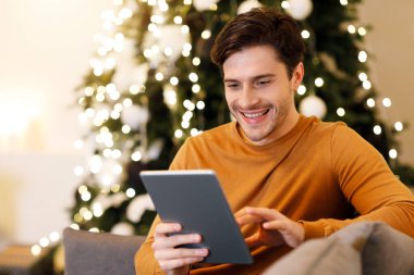 People And Technology. Portrait Of Smiling Handsome Man Holding And Using Digital Tablet Sitting On Couch Indoors In Living Room. Happy Male Surfing Internet, Watching Movie Online, Choosing Gift