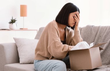 Yanlış madde kavramı. Üzgün Asyalı kadın koltukta oturuyor ve karton kutuları açıyor. Zarar görmüş personelle dolu bir paket. Hayal kırıklığıyla yüzünü kapatıyor.