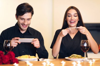 Portrait of excited young millennial couple sitting in restaurant and taking pictures of food with mobile phones for social media, foodie bloggers celebrating Valentines Day together, recording video