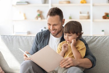 Hikaye zamanı. Sevgi dolu bir baba küçük oğluna ilginç masallar okuyor, ilgili sevimli bir çocuk babasıyla eğleniyor, evdeki kanepede birlikte oturuyor.