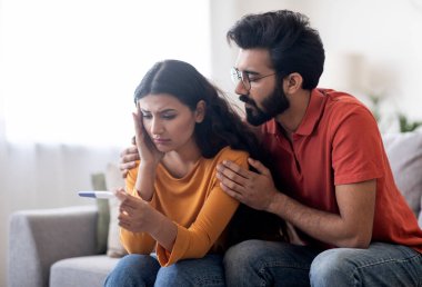 Negatif gebelik testi olan üzgün Hintli çift evdeki koltukta oturup sonuçlara bakıyorlar, depresyonda olan doğulu erkek ve kadın kısırlık sorunu yaşıyorlar.
