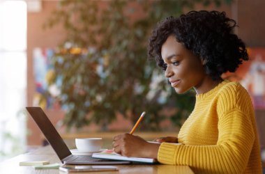 Genç siyahi bayan dizüstü bilgisayarın önünde not alıyor, kafede iş arıyor, boş bir alanda.